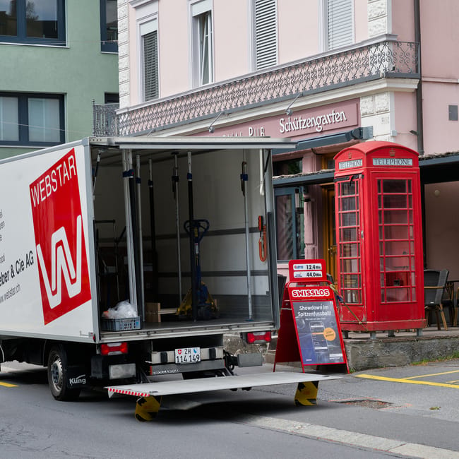 A white moving van with the logo 'WEBSTAR' parked on a street in Belgravia, with its rear door open revealing an empty interior equipped with a hand truck, a small blue plant lift, and packing materia