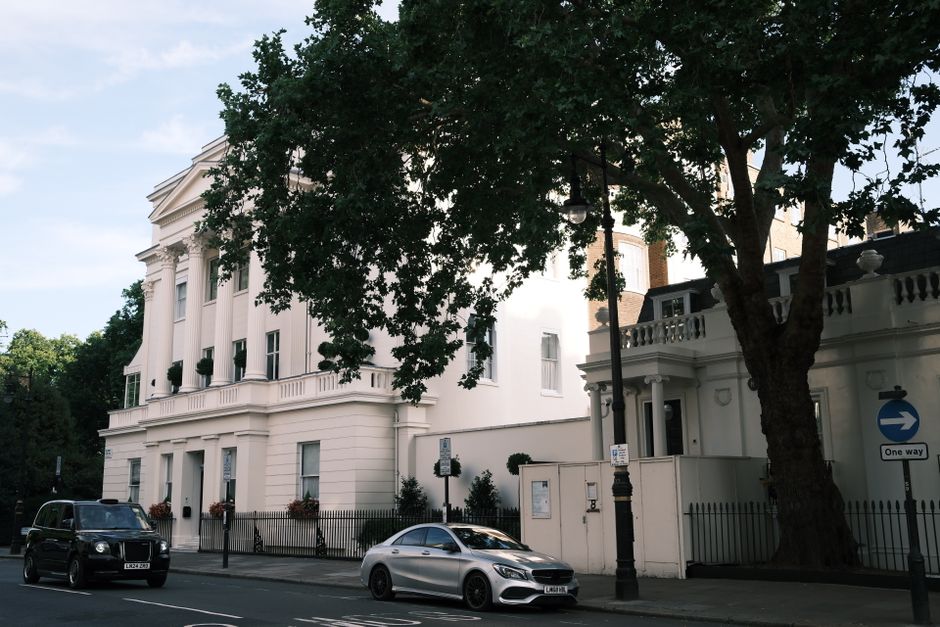 A street scene outside a large, white, classical-style building in Belgravia with tall windows, decorative columns, and a small fenced garden area with potted plants. Two cars, a black van and a silver sedan, are parked along the curb in front of the property. A black lamppost and a one-way street sign are visible on the pavement. In the foreground, a large tree with dark green leaves partially obscures the building, casting shade over the scene during daylight. This setting reflects a residential area suitable for home relocation and moving services, with clear visibility of property access and transport vehicles involved in furniture transport and packing and moving activities, as managed by Man With a Van Belgravia.