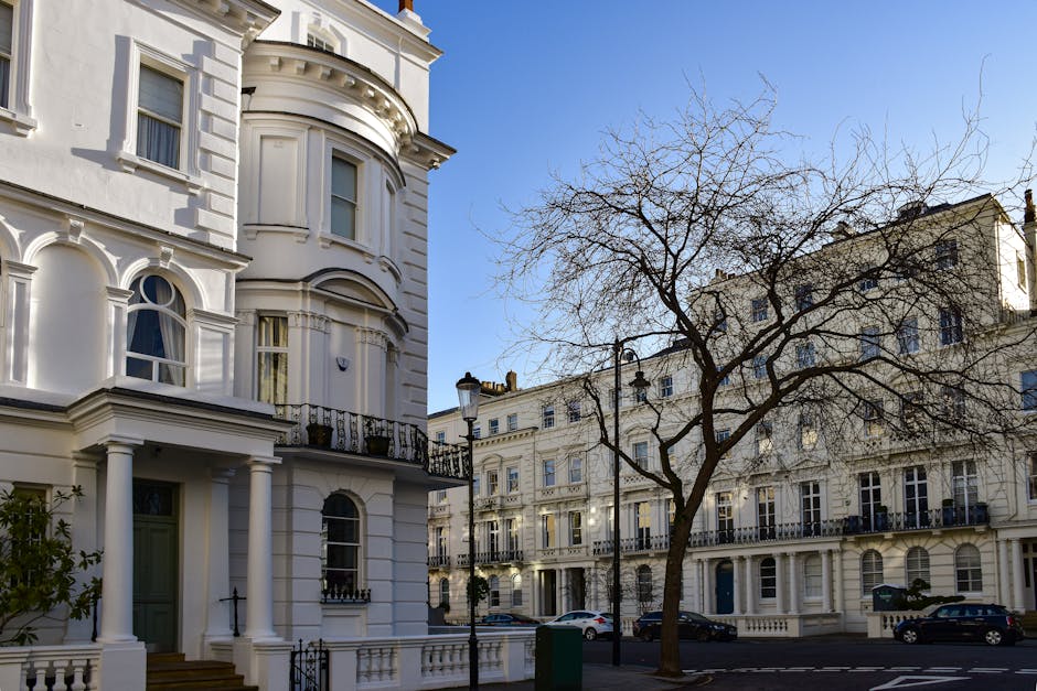 A multi-storey red brick residential building with ornate architectural details, including arched windows and decorative stonework, located in Belgravia. The building features several balconies with metal railings, and the ground floor has a central entrance with steps leading up to two white doors. In front of the building, there is a paved pavement with a double yellow line marking the roadside. The sky above is clear and light blue, indicating daytime. The scene appears to be part of a house removal or home relocation process, typical of services offered by Man With a Van Belgravia, with the building awaiting or undergoing furniture transport. The overall setting reflects an urban residential area with a historical Victorian or Georgian façade, suitable for professional removals and packing services related to house moves in Belgravia.