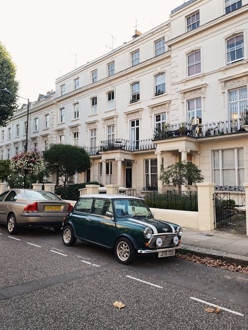 A classic green Mini Cooper with a white roof and racing stripes is parked on a residential street in front of a row of white, multi-storey terraced houses with balconies and large sash windows. Next to the Mini, there is a modern grey hatchback car. The street is lined with trees and small hedges, with some flowering plants visible near the front gardens of the houses. The pavement is clean, and the road has white dashed lines marking parking spaces. In the background, the buildings are well-maintained with decorative moldings around the windows and iron railings on the balconies. The scene is set during daylight hours with overcast weather, providing natural lighting that highlights the architecture and parked vehicles. This setting reflects typical urban residential surroundings suitable for home relocation or furniture transport, with the presence of cars indicating preparation for moving logistics, as might be coordinated by a professional removals company such as Man With a Van Belgravia.