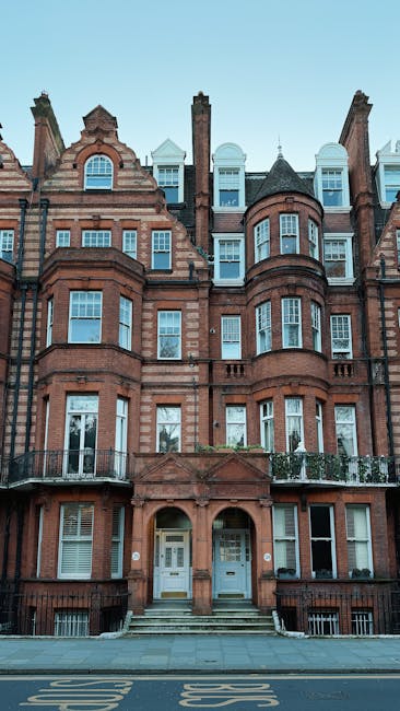 A row of elegant, white Victorian-style townhouses with ornate architectural details, large windows, and decorative mouldings, situated on a wide residential street in Belgravia. The street is lined with leafless trees with pruned branches, casting shadows on the pavement. Several parked cars of various sizes and colors occupy designated parking spots along the curb. The scene is captured in daytime with clear blue sky and some scattered clouds, indicating fair weather. The environment reflects a quiet, affluent neighbourhood typical for house removals and relocations in London, with no visible moving activity in progress but consistent with a setting where professional relocation services, such as those provided by Man With a Van Belgravia, might operate.