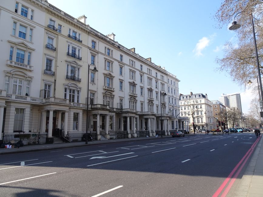 A wide view of a row of elegant, multi-storey residential buildings in Belgravia, London, with classical architectural features including tall windows, decorative balconies, and stone facades. The buildings are situated along a paved street with marked parking spaces and a red curb, under a clear blue sky with some scattered clouds. On the street, several parked cars can be seen, and on the pavement, there are stacks of cardboard boxes, padding, and wrapping materials indicating preparations for a home relocation. A person is visible in the distance near a black van, possibly engaged in a loading process, with the van parked close to the building's entrance. The scene suggests a professional moving and furniture transport activity coordinated by Man With a Van Belgravia, requiring careful handling of furniture and boxes, and reflecting the logistical aspects of packing and moving services in the Belgravia area.