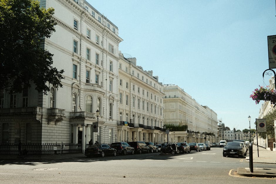 A street scene in Belgravia showing a multi-storey residential building with brown brick facades, large sash windows, and small balconies. The building is situated directly adjacent to the pavement, which has metal bike racks and a bicycle parked in front. Several delivery trucks and cargo vans are parked along the street, some positioned close to the building’s entrance for loading and unloading. Visible in the foreground are cardboard boxes, wrapped furniture pieces, and protective blankets indicating a home relocation process. Few people are walking along the pavement, and the overcast sky creates diffuse natural light. This scene captures the typical environment for furniture transport and packing during house removals managed by [COMPANY_NAME], operating within London’s Belgravia area, as part of a professional removals service focused on efficient moving logistics and loading procedures.